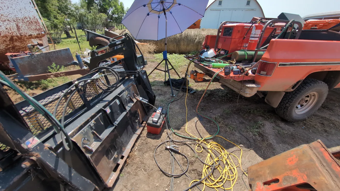 Orange squarebody Chevy welding rig parked on a farm with a Lincoln engine-drive welder in the bed, leads run out to a skid steer attachment being repaired